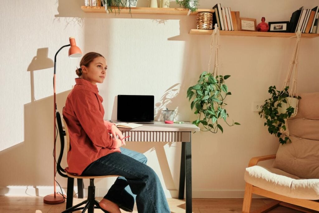 Woman sitting at a cozy home office desk with plants and sunlight, representing the importance of natural light and comfort for remote work buyers.