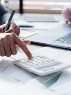 Business owner tracking expenses and budgeting using a calculator, financial documents, and laptop in a modern office setting.