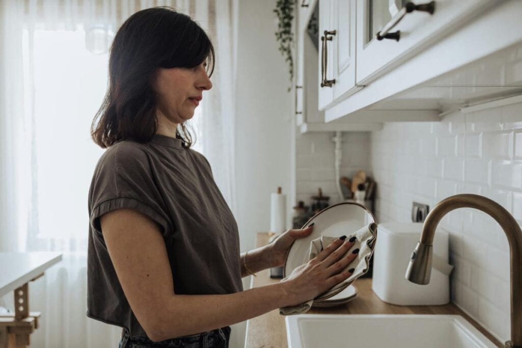 Woman stands at the kitchen sink drying a plate with a cloth, surrounded by clean white cabinetry and soft natural light