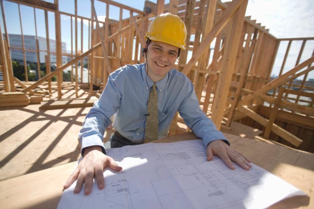 Construction manager reviewing building plans at a job site with wood framing in the background.