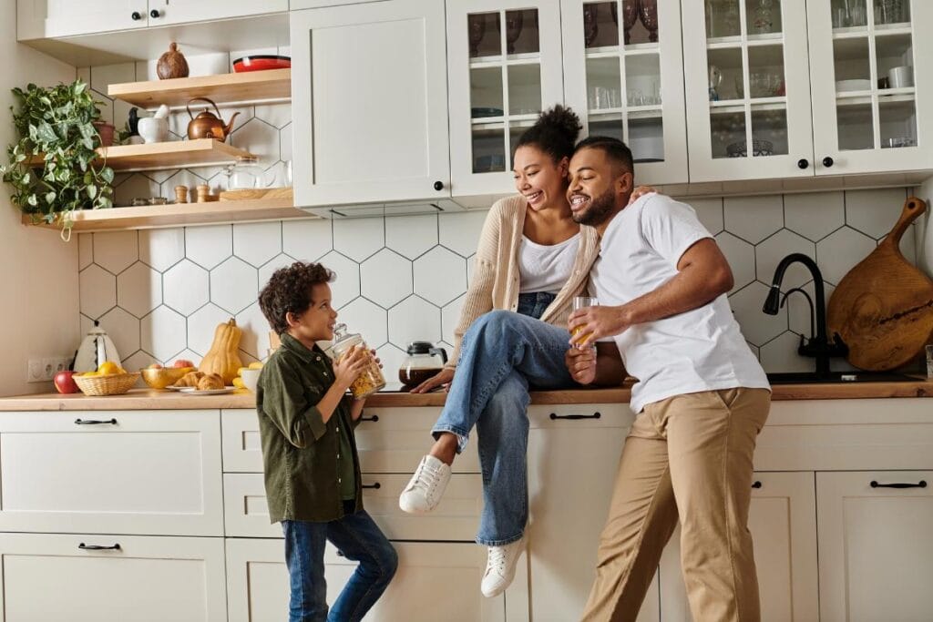 A smiling family spending time together in a bright modern kitchen with white cabinets, hexagon tile backsplash, and open shelving.