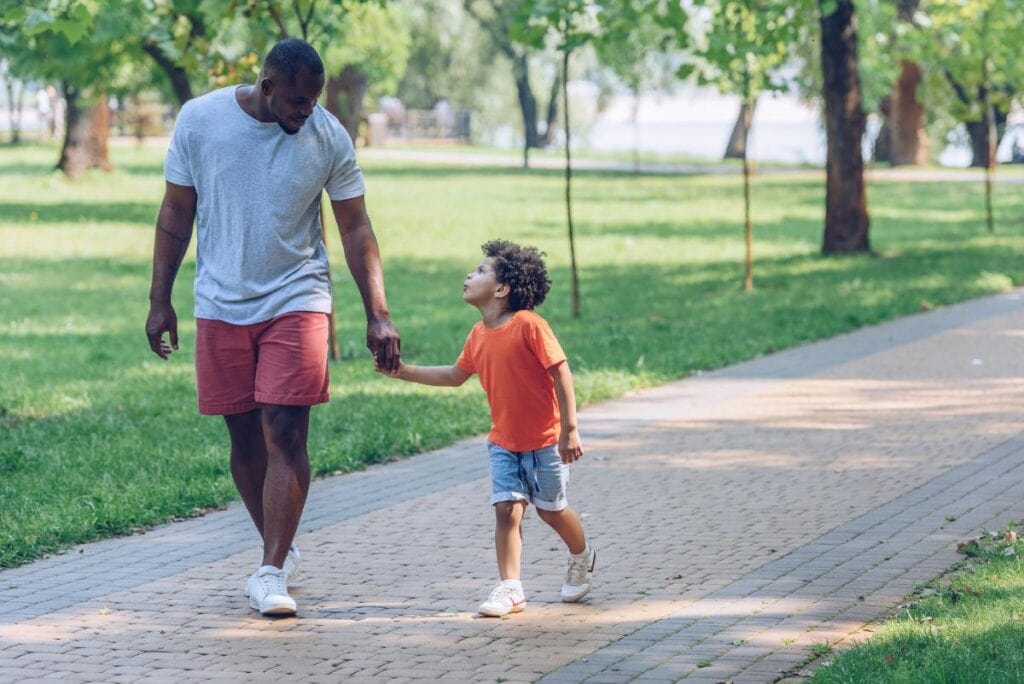 Father and young son holding hands and walking together on a tree-lined park path, enjoying a peaceful nature walk.