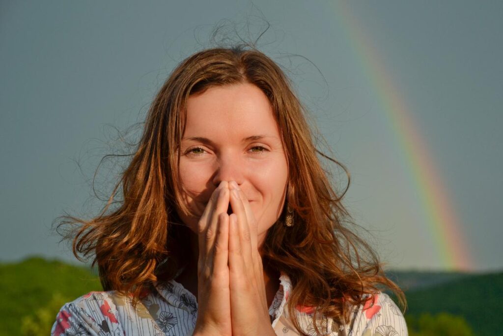 Smiling woman holding her hands near her face outdoors with a rainbow in the background, symbolizing gratitude and joy.