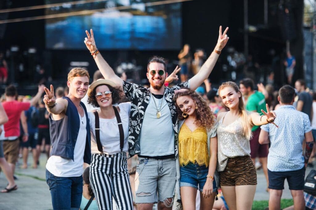 A group of young friends smiling and posing with peace signs at an outdoor concert, excited for the show to begin.
