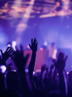 Silhouetted concert crowd with hands raised in the air under bright purple stage lights.