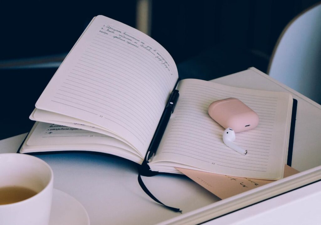 An open journal with a pen, wireless earbuds, and a cup of tea on a desk, representing journaling and mindfulness habits