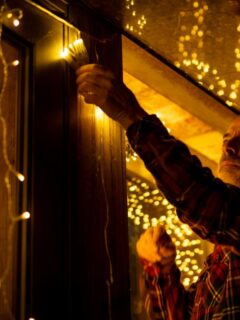 A man in a plaid shirt carefully installs warm white Christmas string lights along a window frame.