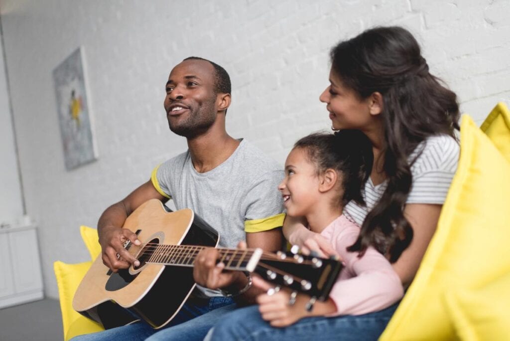 Father playing acoustic guitar with daughter and mother smiling beside him on a couch, sharing a joyful musical moment.