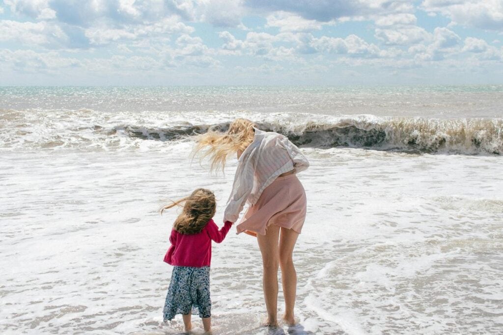 Mother and daughter holding hands while standing at the edge of the ocean, enjoying a breezy, carefree moment together at the beach.