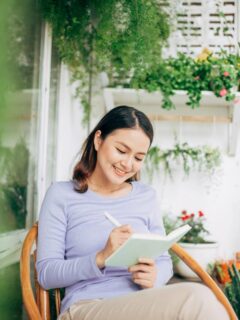 Woman sitting on a balcony surrounded by plants, smiling while writing in a journal, capturing a peaceful and joyful moment.