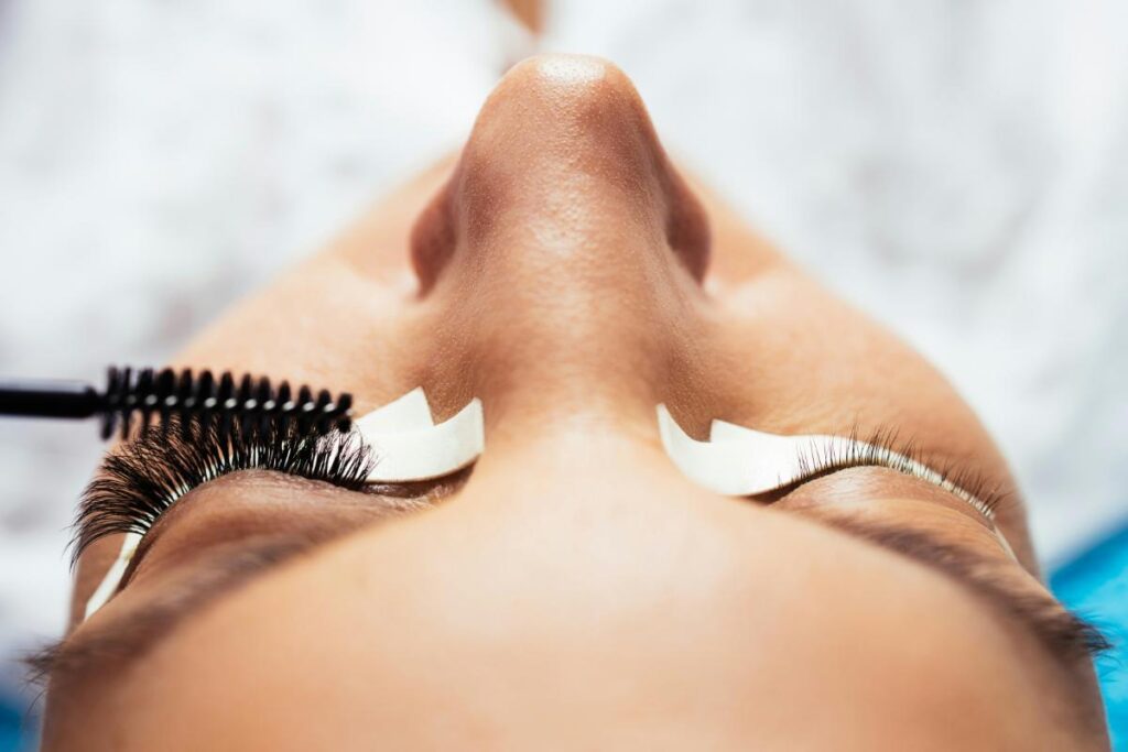 Lash artist brushing newly applied premade volume fans on a client's closed eye, showing the difference between natural and extended lashes.
