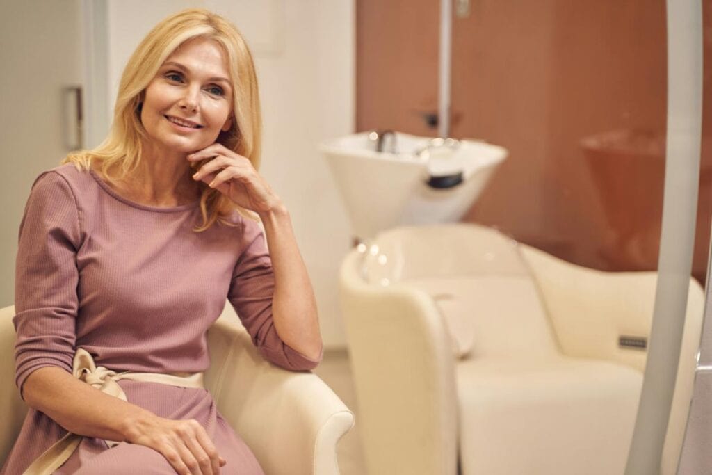 older blonde in pink or mauve dress sitting in a salon
