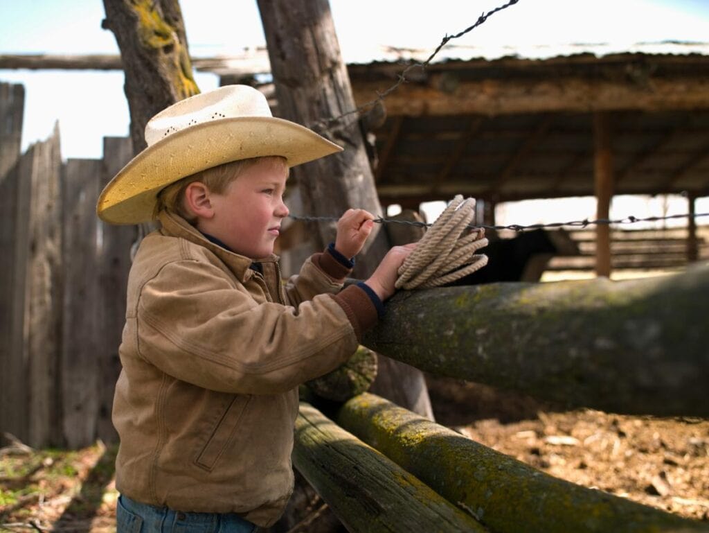 Young boy in a cowboy hat and tan jacket holding a rope and looking through a wooden fence on a ranch.
