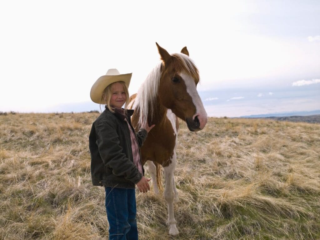 Smiling girl in a cowboy hat and jeans standing next to a brown and white horse in an open field.