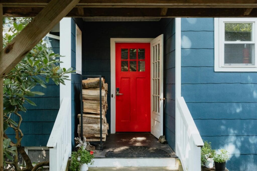 Front porch of a home with a bold red entry door framed by white trim, featuring stacked firewood and plants, enhancing curb appeal and character.