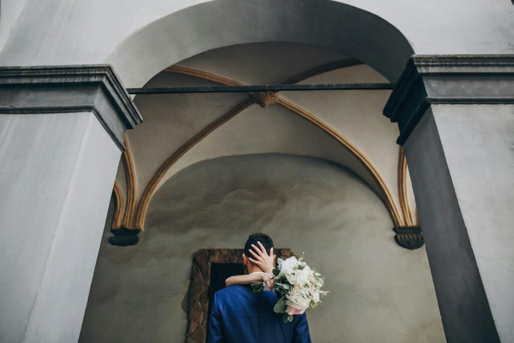 A bride embraces a groom under an elegant arched ceiling, holding a bouquet of white and blush flowers behind his back