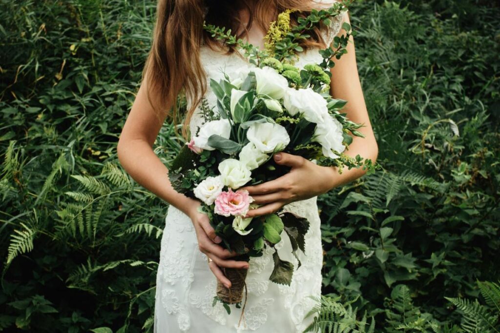 A bride in a lace dress holds a rustic bouquet of white, green, and pink flowers while standing amidst lush greenery.