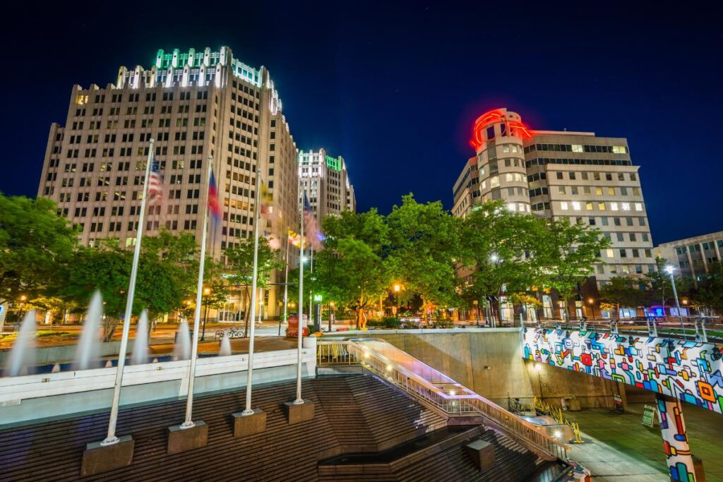 Night view of downtown Bethesda, Maryland featuring tall office buildings with colorful rooftop lights, waving flags, a modern fountain, and public art near the Metro station.