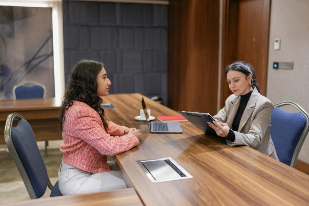 A young woman sits across from an interviewer at a wooden conference table, attentively listening as the interviewer reviews a clipboard.