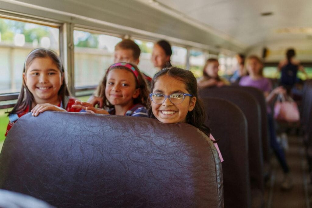 Smiling children sitting on a school bus, excited for a field trip, with chaperones and other students in the background.