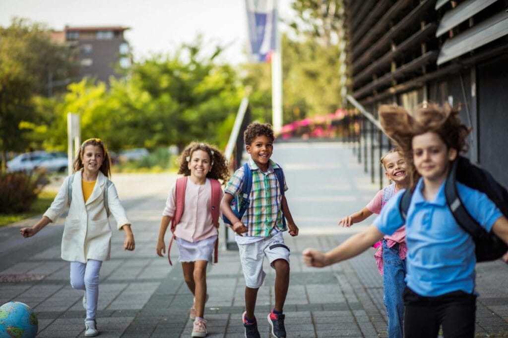 Group of young children happily walking together outside a school building, wearing backpacks and smiling.