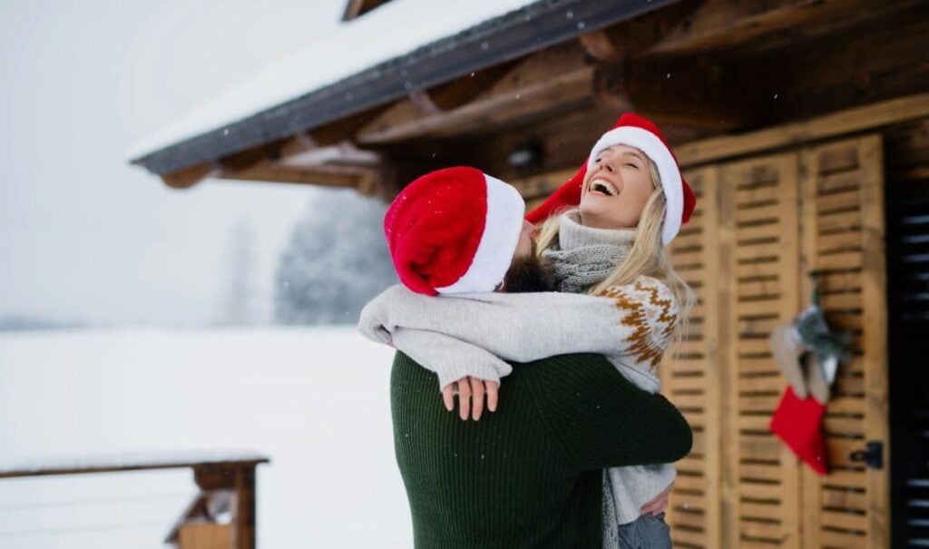 A couple wearing Santa hats shares a joyful hug outside a snow-covered cabin, surrounded by a festive winter setting.