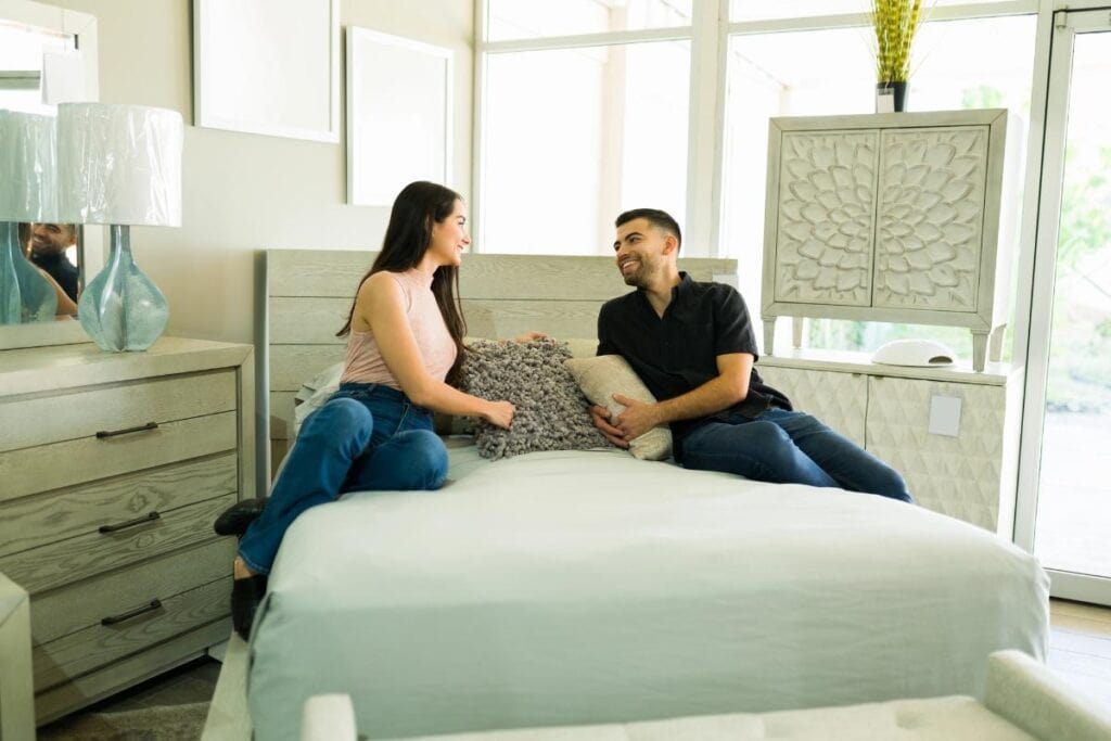 A smiling couple sits on a bed in a bright furniture showroom, discussing and testing the comfort of the mattress and pillows.