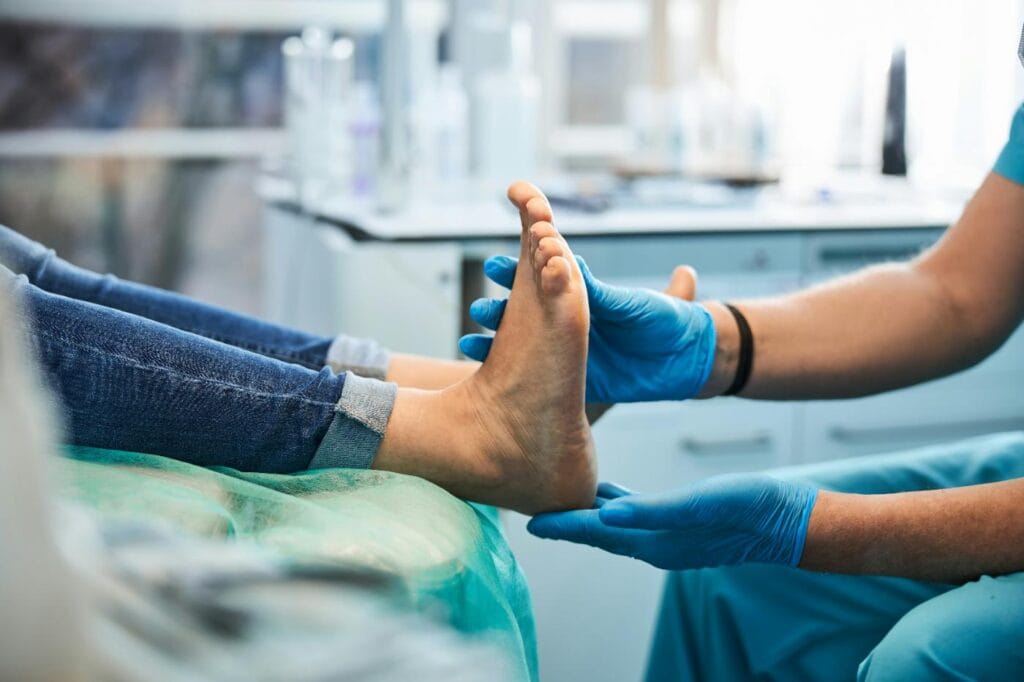 A podiatrist wearing blue gloves examines the bottom of a patient's foot during a consultation, with the patient lying on an examination table.