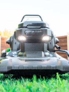 Front-facing close-up of a modern black electric lawn mower with LED lights, sitting on freshly cut grass in a fenced backyard.