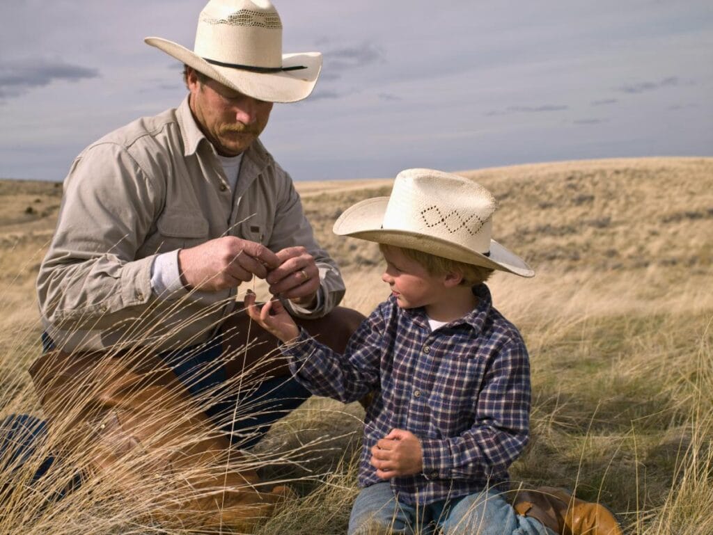 Father and young son in cowboy hats and plaid shirts sitting in tall grass, focused on a hands-on outdoor task.