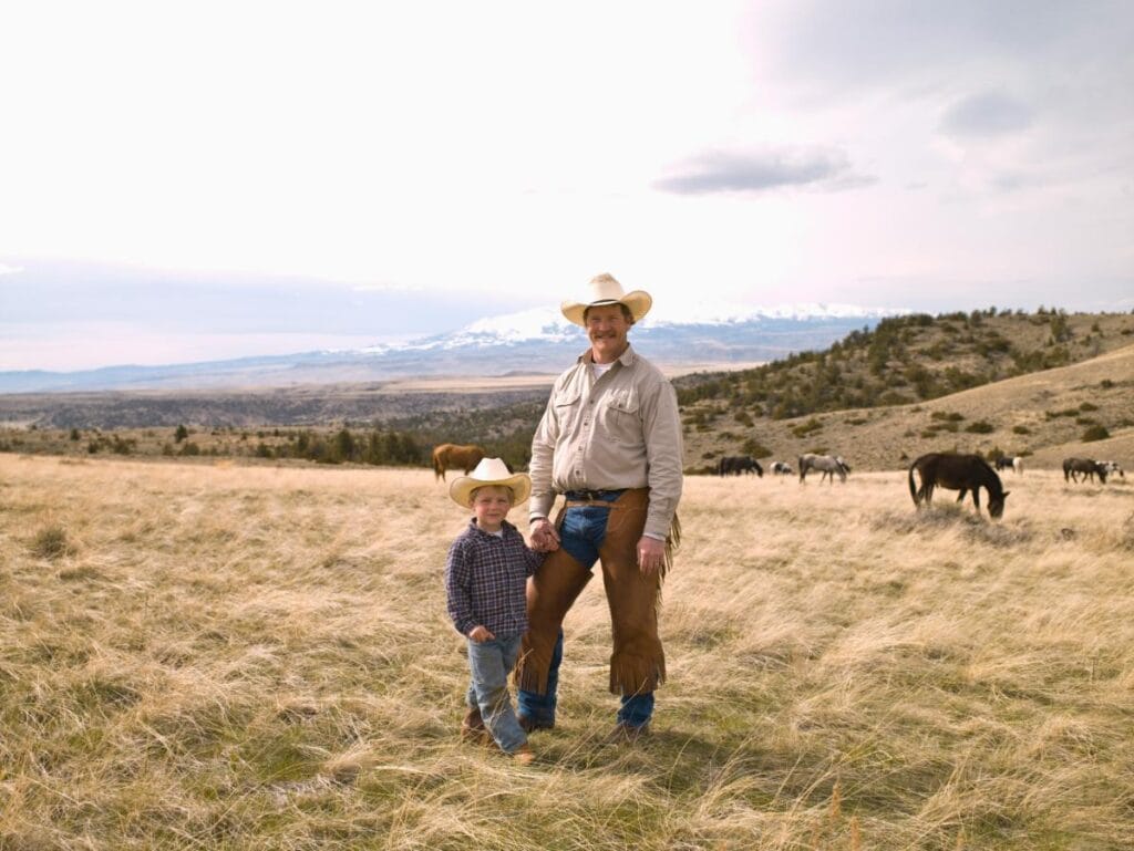 Cowboy father and son standing together in a wide open field with grazing horses and distant mountains behind them.