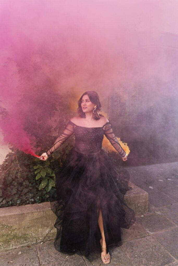 A woman in a black formal gown holds smoke bombs emitting pink and yellow clouds, creating a bold and atmospheric scene during an outdoor event.