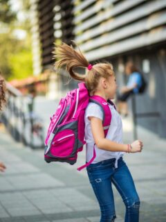 Excited schoolchildren running and playing near a school, with backpacks bouncing and laughter visible on their faces.