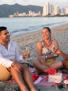 Three friends celebrate Christmas on the beach, enjoying drinks and laughter while sitting on a picnic blanket with gifts and decorations.