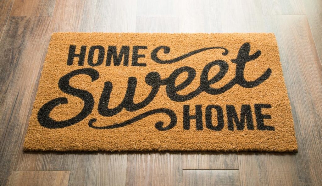 A rectangular coir doormat with the phrase "Home Sweet Home" in bold, decorative lettering on a wood floor entryway.