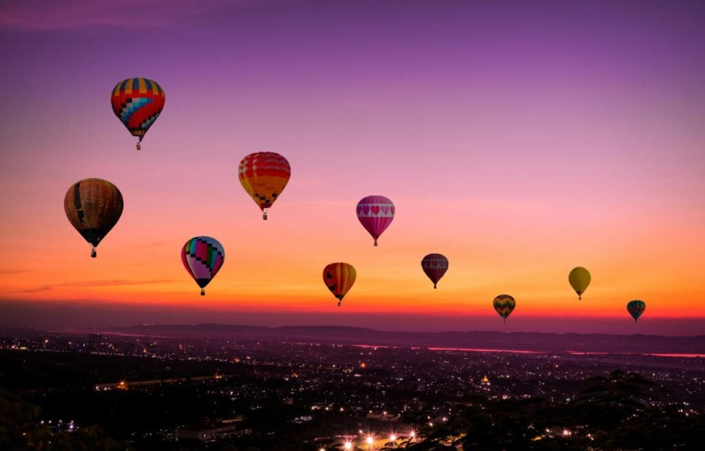 Multiple colorful hot air balloons drift peacefully across a vivid sunset sky over a cityscape, creating a magical and serene scene.