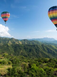 Two vibrant hot air balloons float above lush green mountains under a bright blue sky, offering a breathtaking view from above.