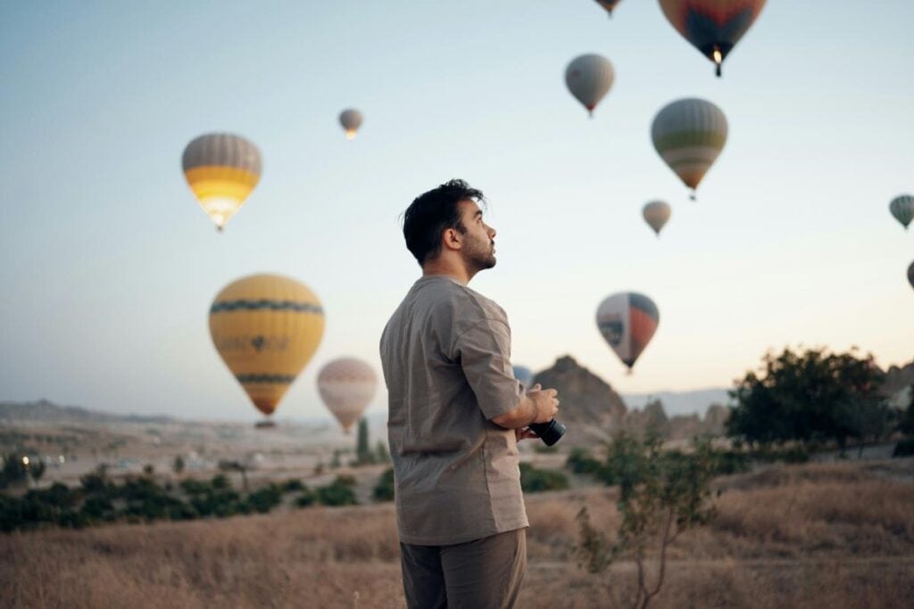 A man stands in a dry field holding a camera, gazing at numerous hot air balloons rising into the soft early morning sky.