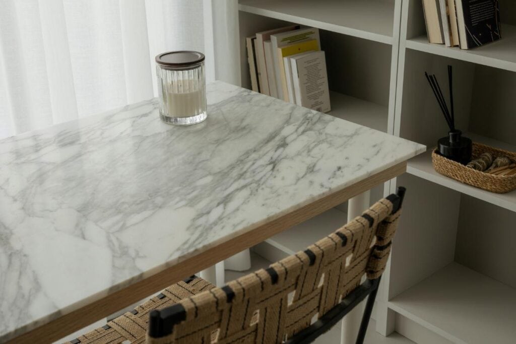 Minimalist home office setup with a marble desk, woven chair, and bookshelves, featuring a glass candle on the tabletop for a clean, elegant look.