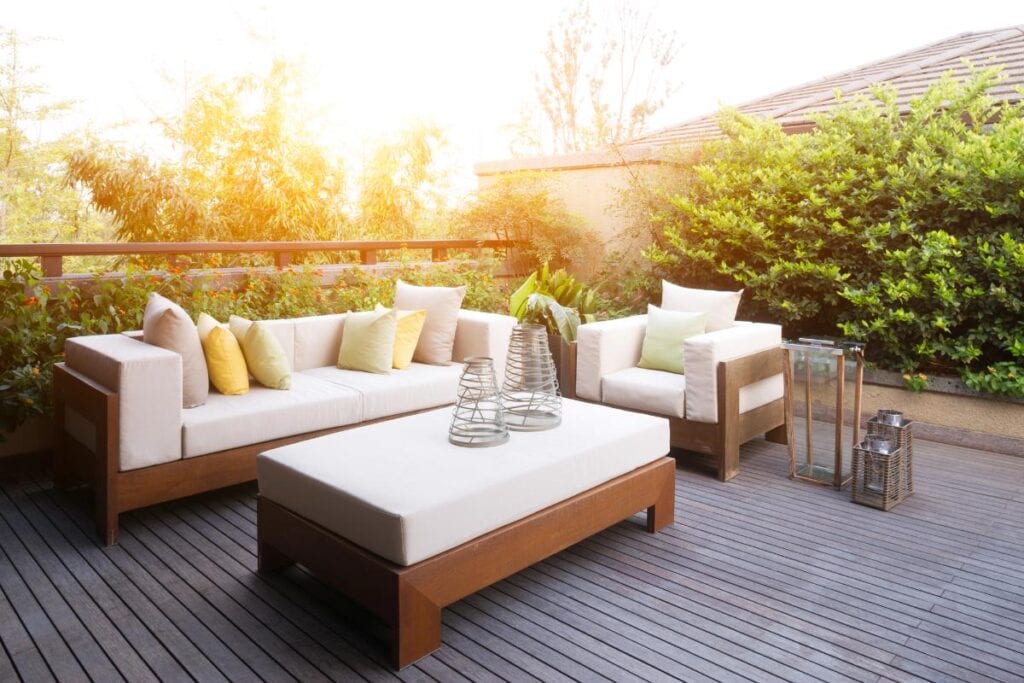 Modern outdoor modular lounge with wooden frame, cream cushions, and pastel throw pillows on a timber deck surrounded by lush greenery at sunset.