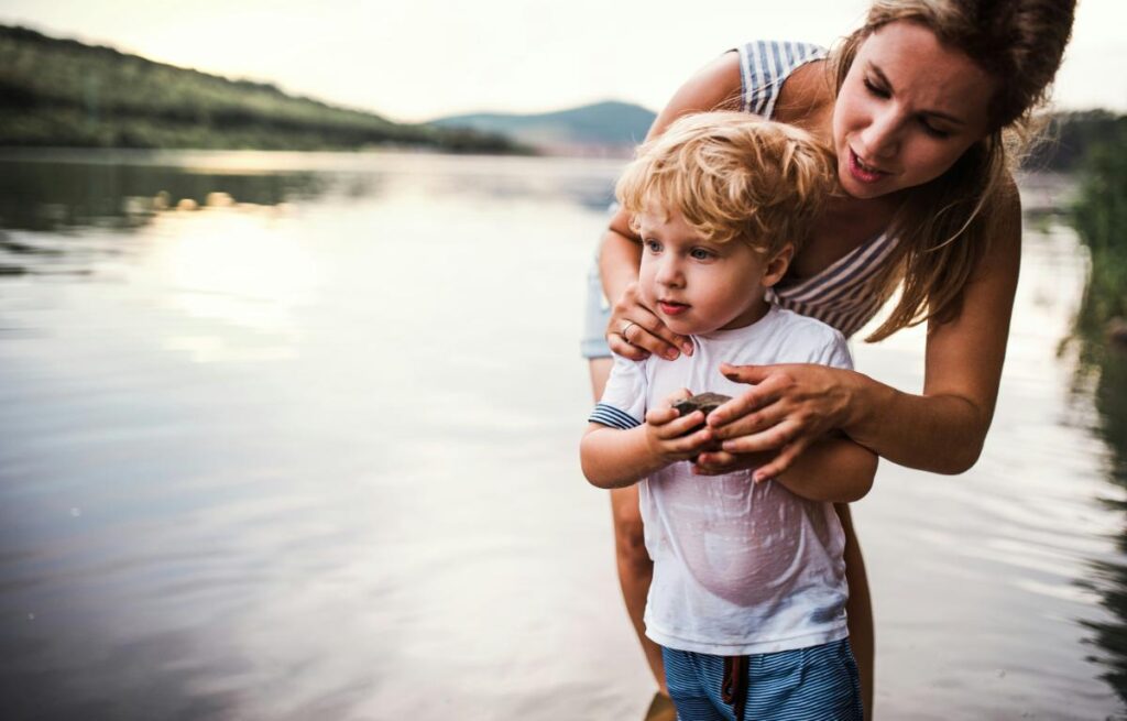 Mother and young boy standing at the edge of a calm lake, closely observing a rock together during a quiet outdoor moment.