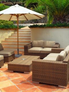 Outdoor modular lounge setting with brown wicker sectional seating, neutral cushions, and patio umbrellas on terracotta tiles surrounded by tropical plants.