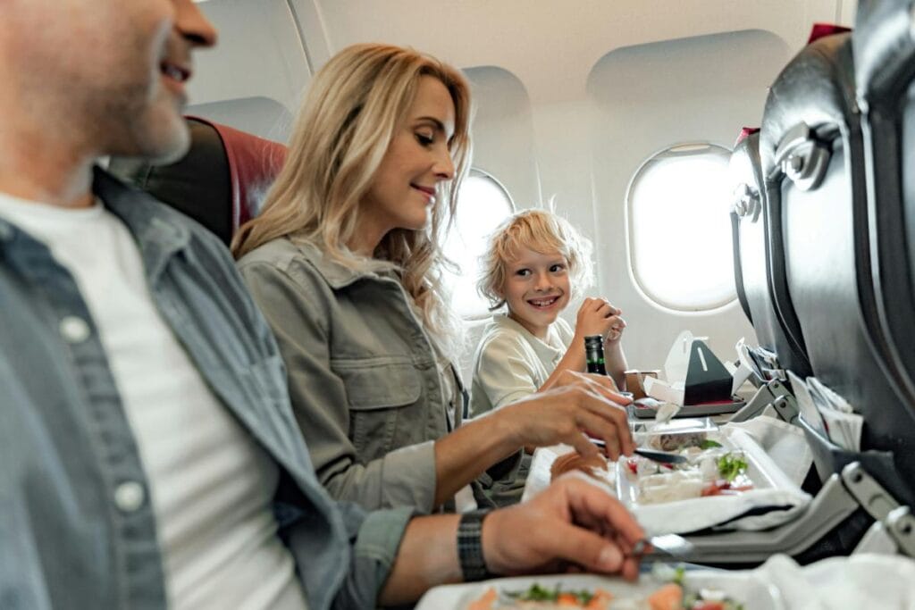 Family seated on an airplane enjoying an in-flight meal, with a smiling child looking at a parent during the dining experience.