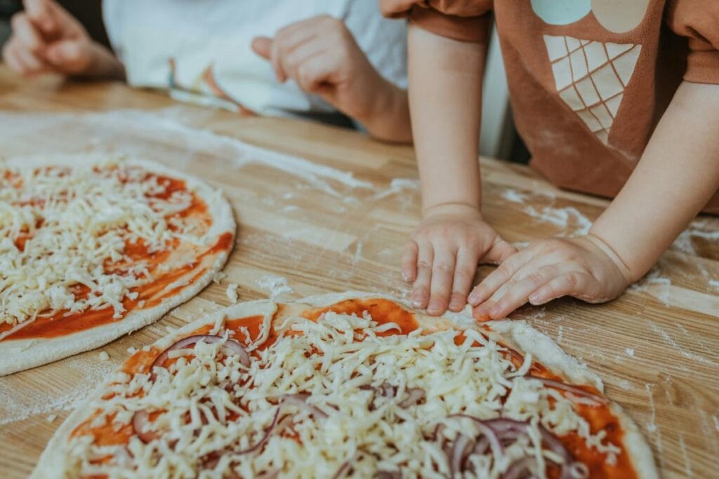 Close-up of young children making homemade pizzas, pressing dough and adding cheese and sauce on a wooden surface, surrounded by flour.