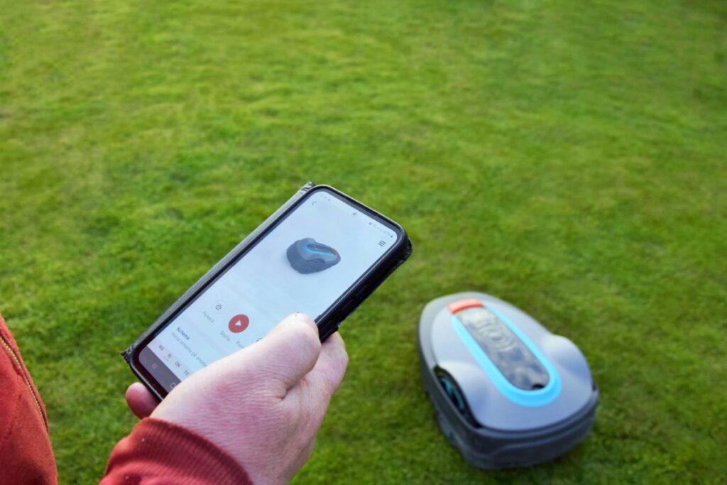 Person using a smartphone app to control a robotic lawn mower on green grass, showcasing smart lawn maintenance technology.