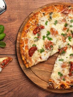 Freshly baked pizza on a wooden board topped with cheese, sun-dried tomatoes, and herbs, next to a cheese grater, Parmesan block, and basil leaves.