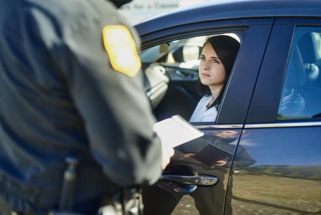 Female driver sitting in a car, looking up at a police officer holding a notepad during a traffic stop in daylight.