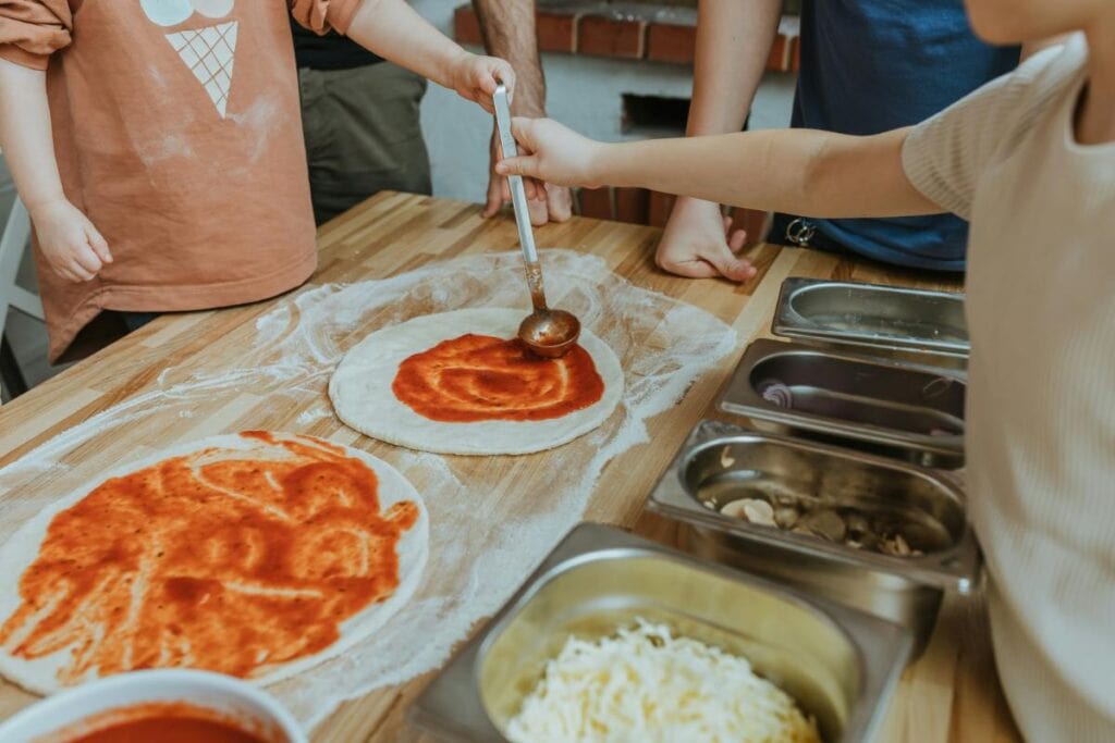 Children spreading tomato sauce on pizza dough using a ladle, with toppings like mushrooms, onions, and cheese ready in stainless steel containers.