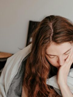 A woman with long hair sits on a bed wrapped in a blanket, resting her head in her hand with a tired, distressed expression.