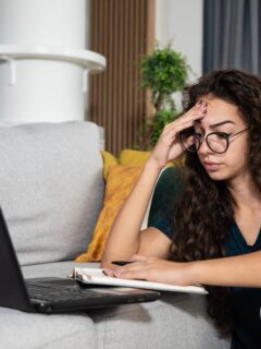 A student sits on a couch with a laptop and notebook, holding her forehead in frustration or fatigue while studying at home.